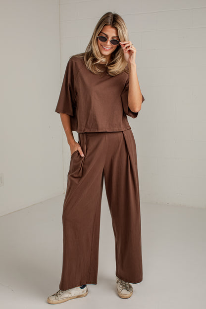 A woman modeling a matching brown two-piece outfit consisting of a short-sleeve cropped top and wide-leg pants, styled with sunglasses and sneakers against a white studio background.