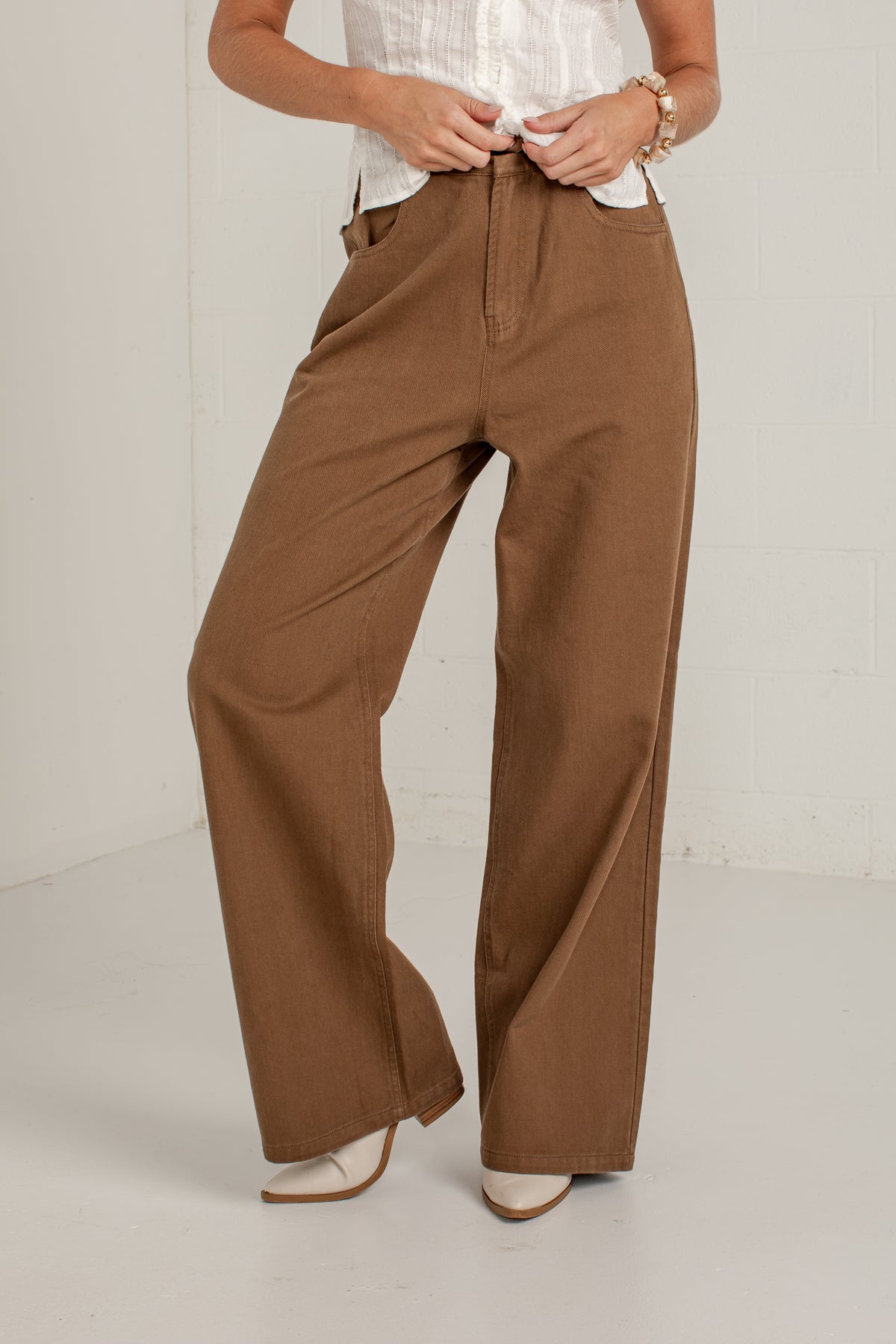 A person wearing high-waisted, wide-leg brown pants with a white top and beige pointed-toe heels, standing against a light gray studio wall.