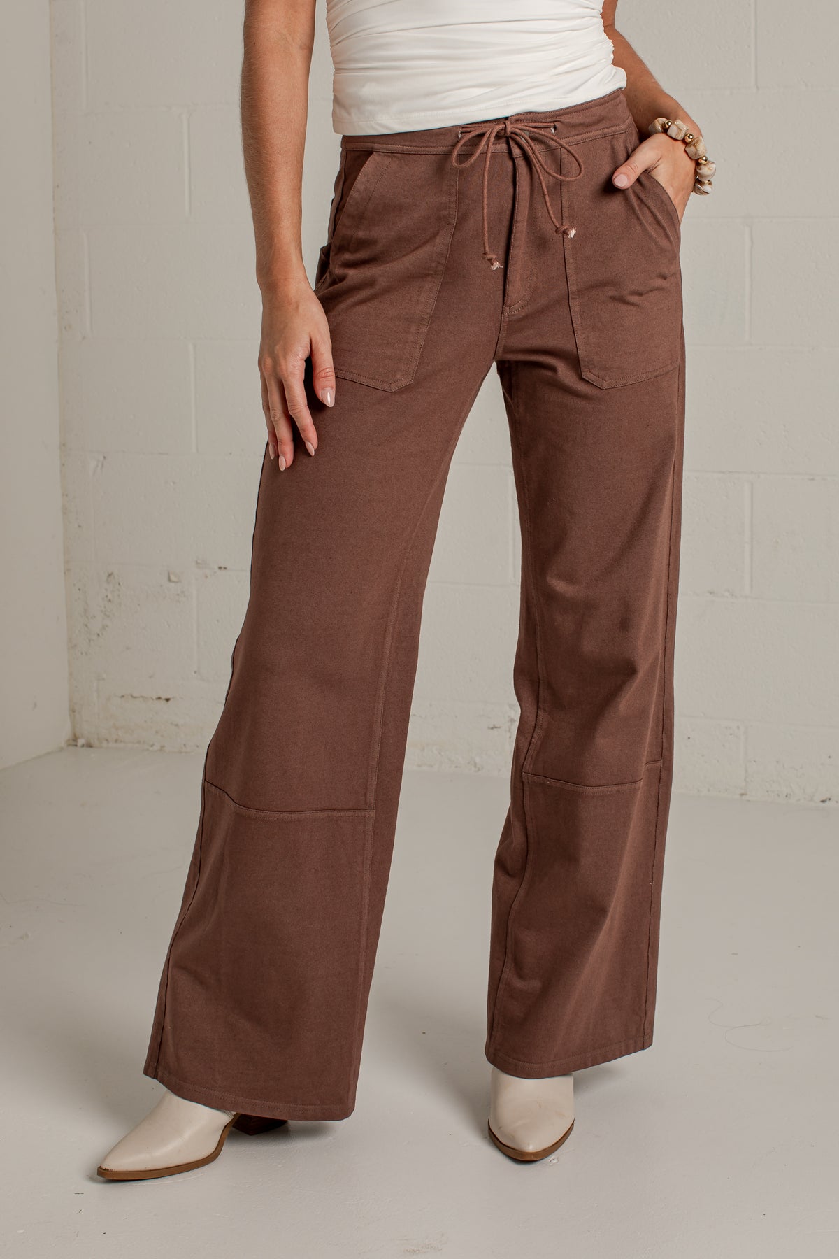 A person wearing brown wide-leg drawstring pants with front patch pockets, paired with a white top and white pointed-toe boots, standing against a light gray wall.