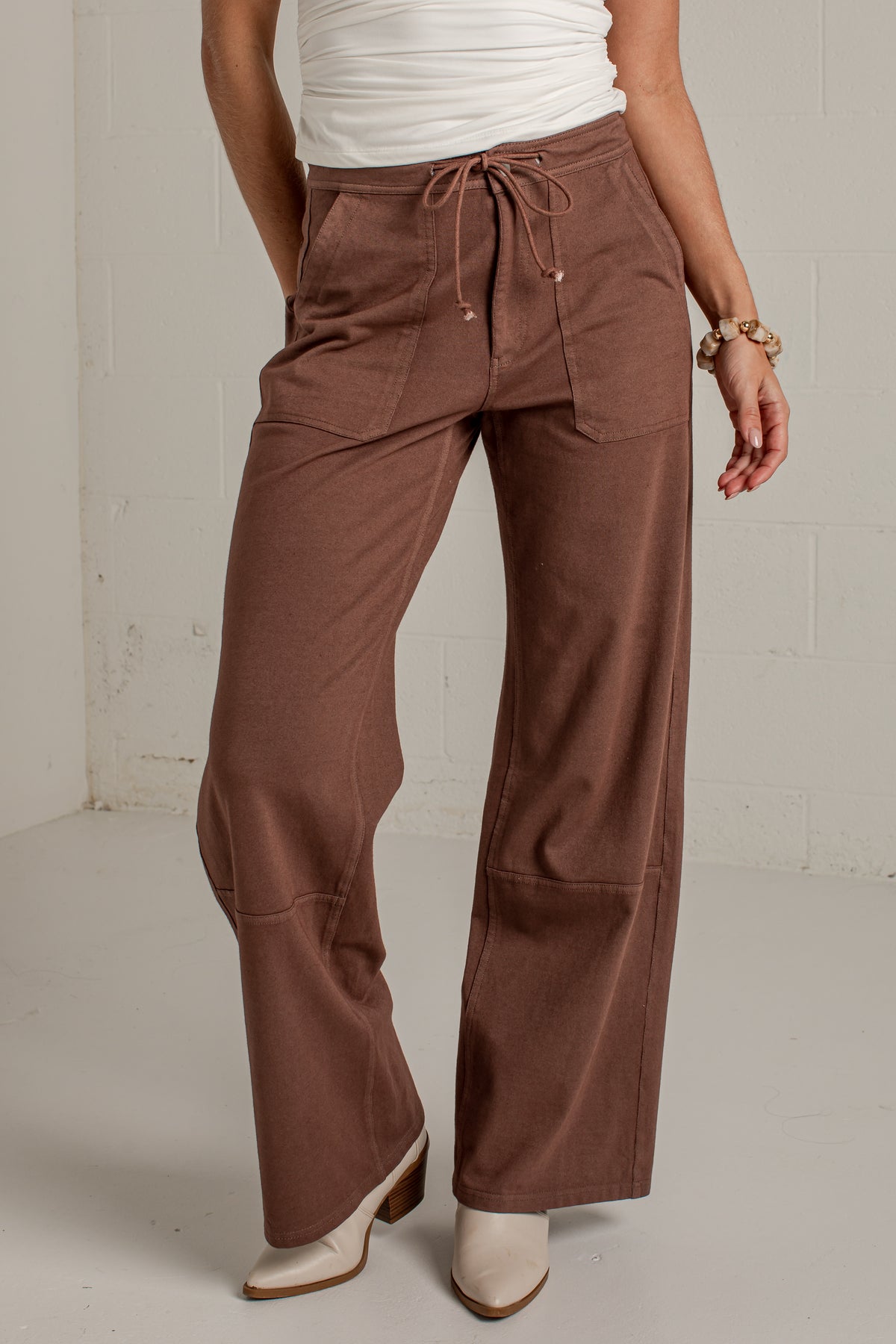A person wearing wide-leg brown pants with a drawstring waist, paired with a white top and white heeled boots, standing against a light gray wall.