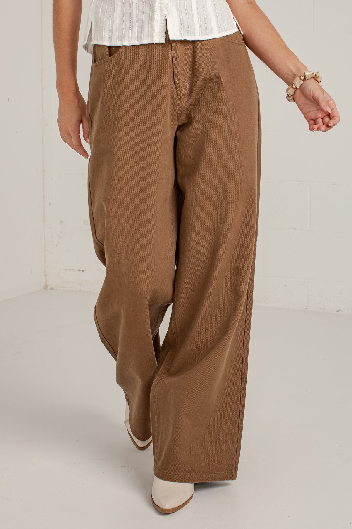 A person wearing wide-leg brown pants, a white top, and white pointed shoes, standing in a bright studio with a white wall and floor.