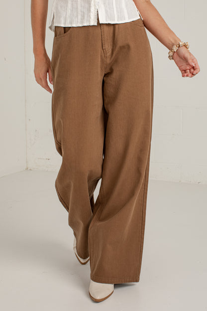 A person wearing wide-leg brown pants, a white top, and white pointed shoes, standing in a bright studio with a white wall and floor.