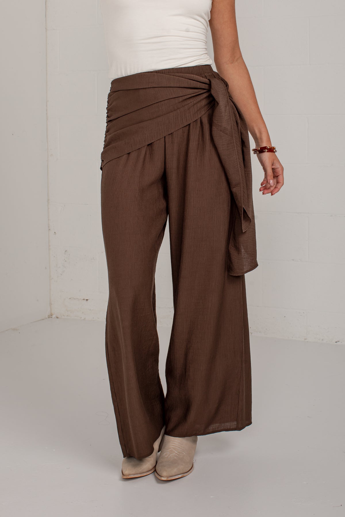 A person wearing brown wide-leg draped pants with a tied waist, paired with a white top and beige ankle boots, standing against a plain light gray wall.