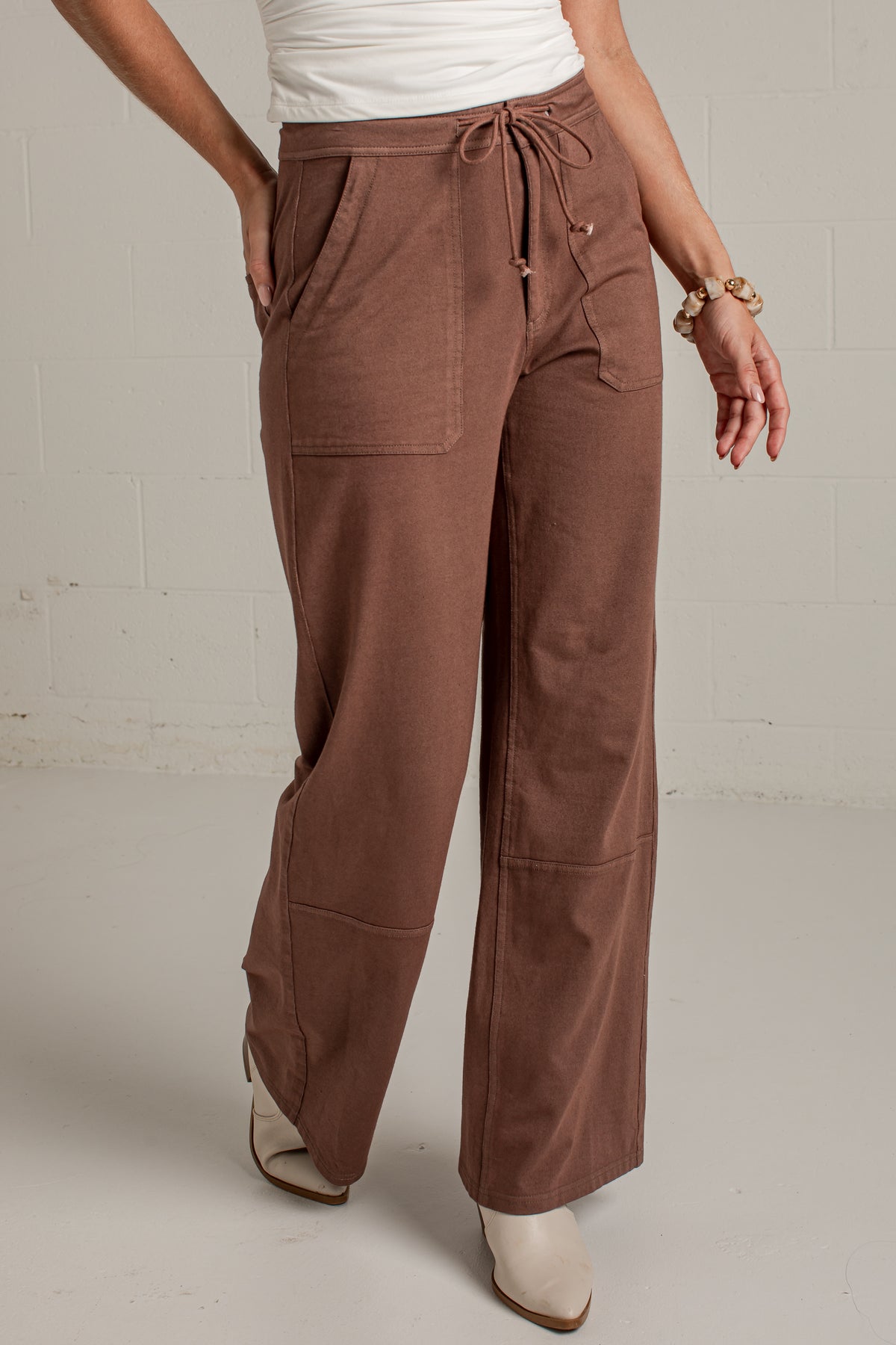 A person wearing wide-leg brown pants with a drawstring waist, side pockets, and patch pockets, paired with a white top and beige pointed-toe heels against a light gray wall.