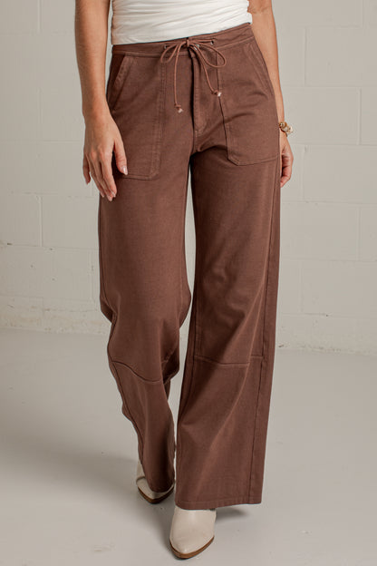 A person wearing wide-leg brown drawstring pants with front patch pockets, paired with a white top and white pointed-toe heels, standing against a light gray wall.