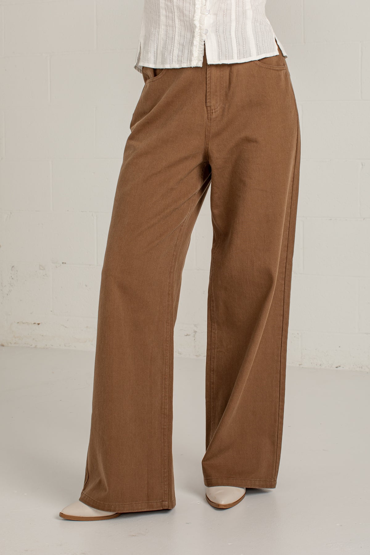 A person wearing brown wide-leg corduroy pants with a white striped top and white pointed-toe flats, standing against a light gray brick wall.