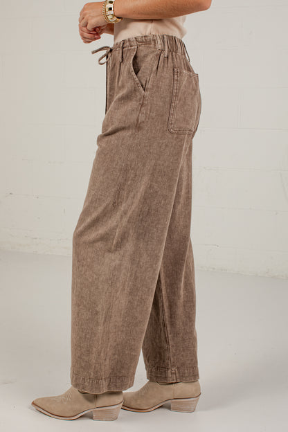 A person shown from the waist down wearing wide-leg brown pants with a drawstring waistband and side pockets, paired with beige ankle boots against a light studio wall.