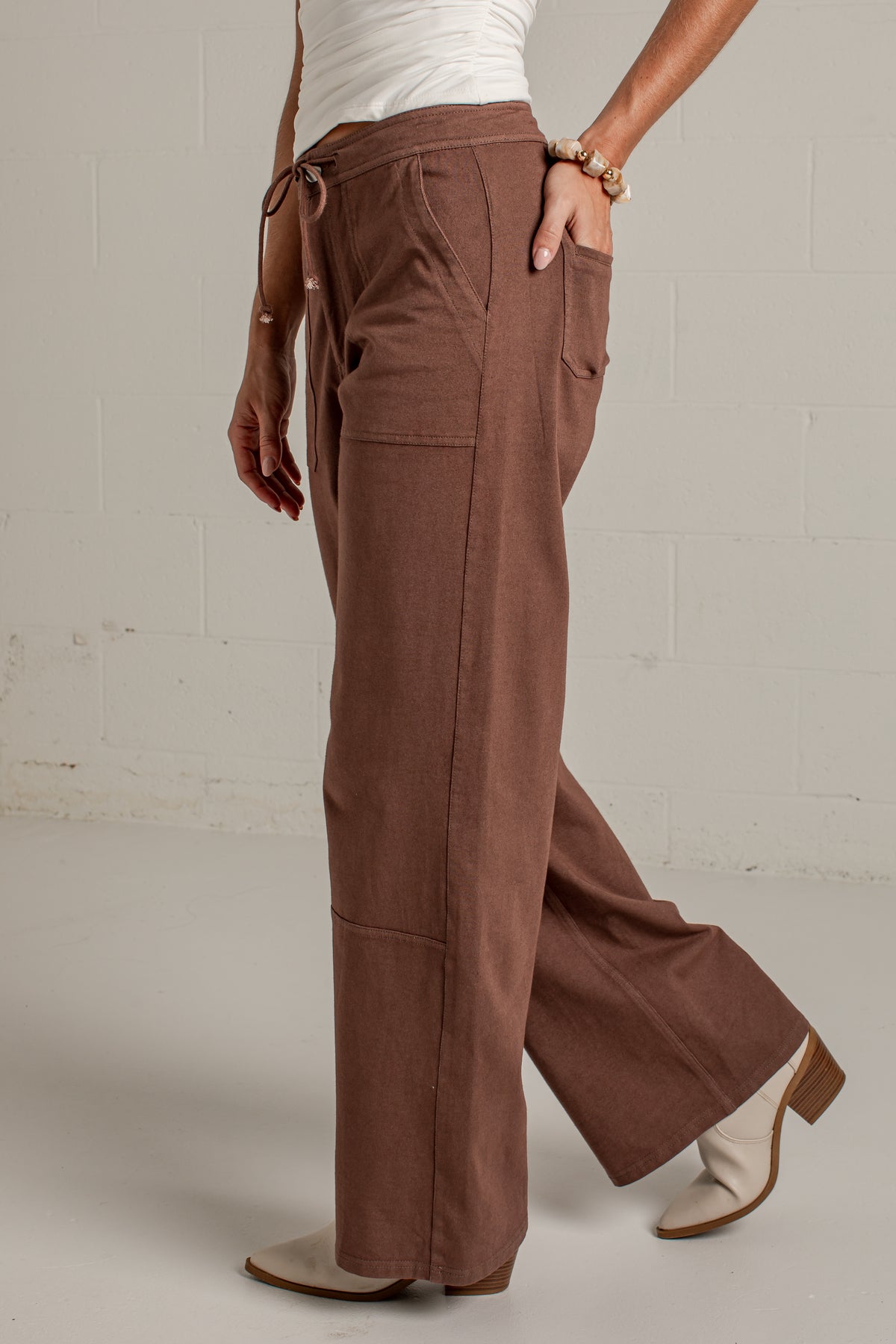 A person wearing wide-leg brown pants with a drawstring waist, shown from the side against a light gray studio wall.
