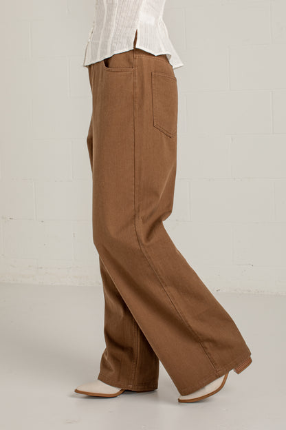 A person wearing wide-leg brown pants with a white blouse and white pointed shoes, shown from the waist down against a light gray brick wall.