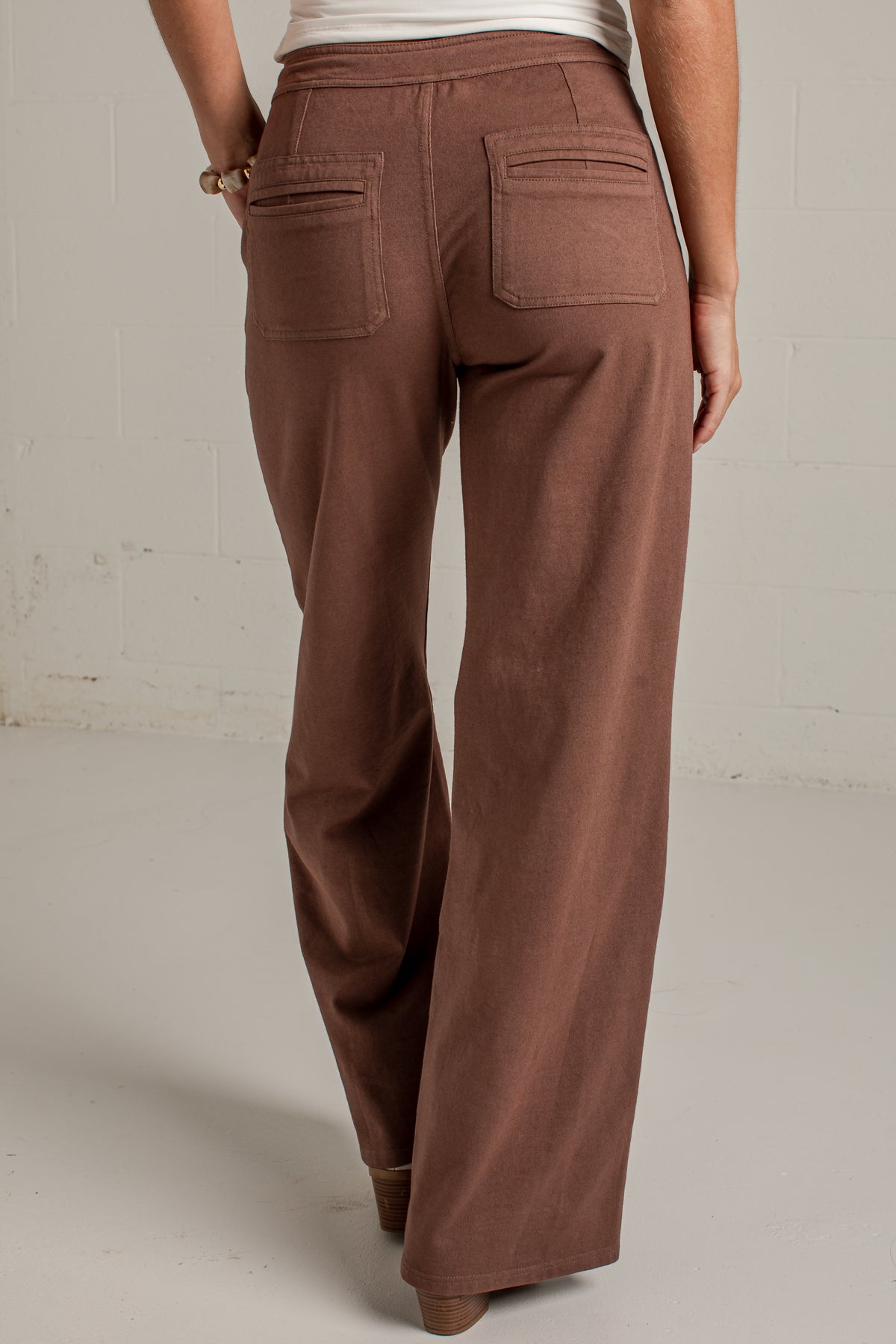 A person shown from the back wearing wide-leg brown pants, photographed in a studio with a light neutral wall and floor.
