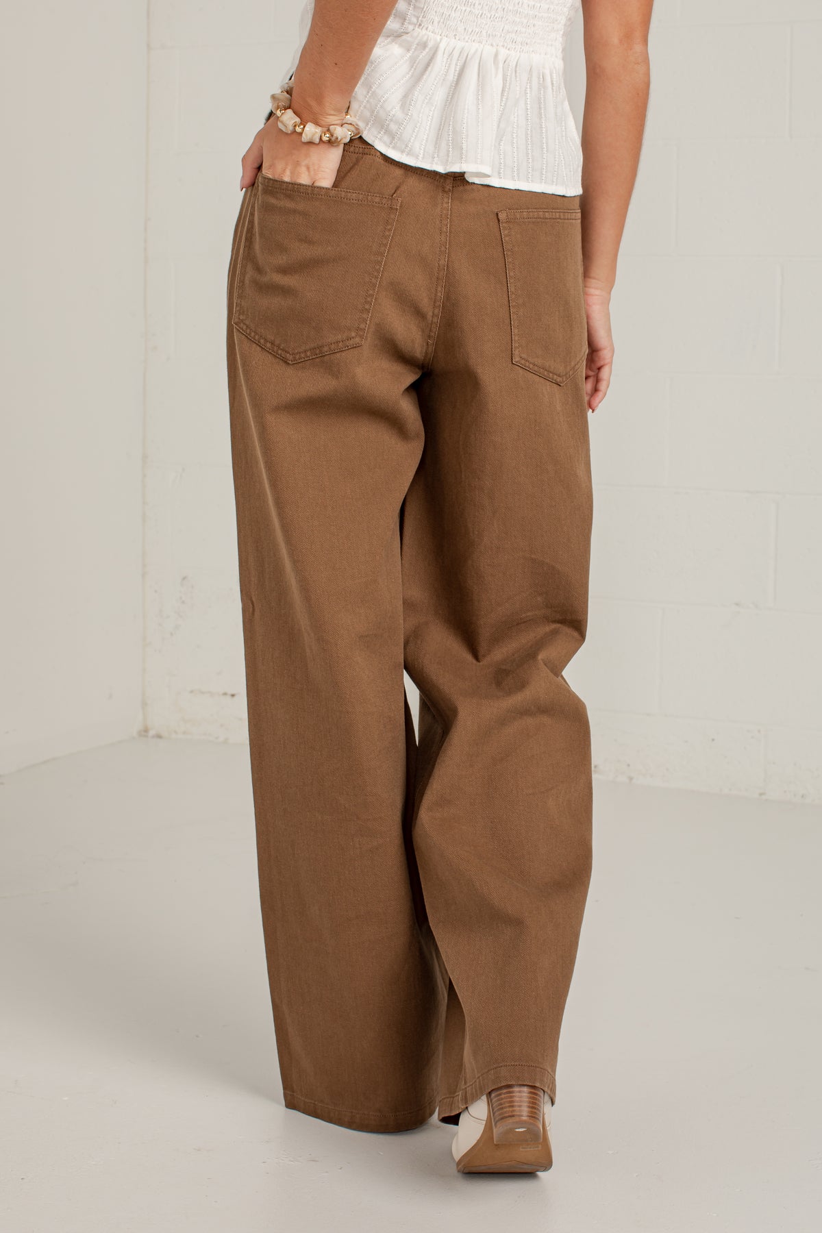 A person wearing wide-leg brown pants with a white ruffled top and brown pointed shoes, standing in a white studio corner.