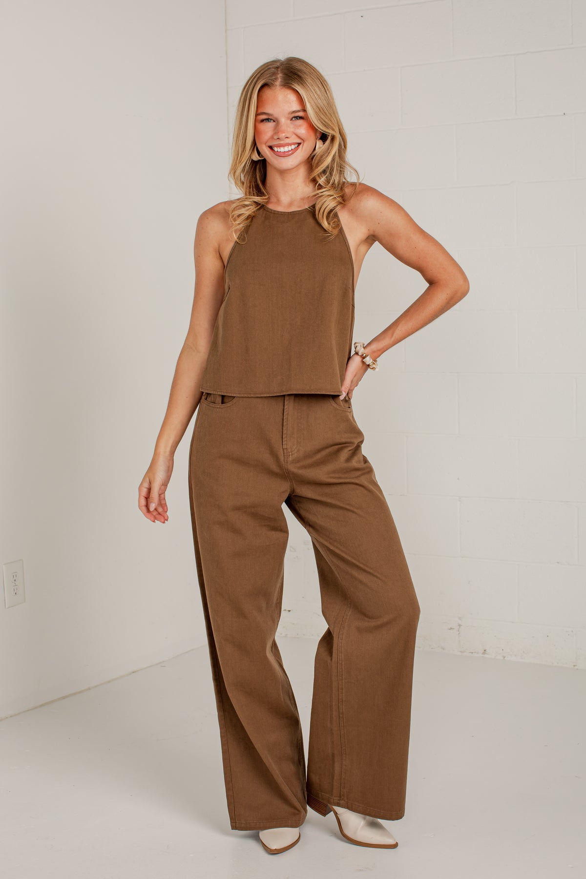 A woman modeling a two-piece brown outfit consisting of a sleeveless halter crop top and matching wide-leg pants, standing against a white studio wall.