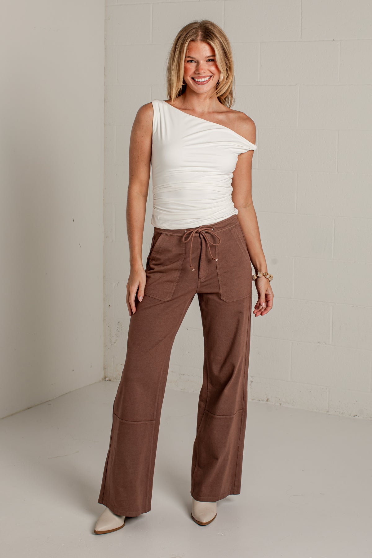 A woman modeling a white one-shoulder ruched top and brown wide-leg drawstring pants in a bright studio setting.