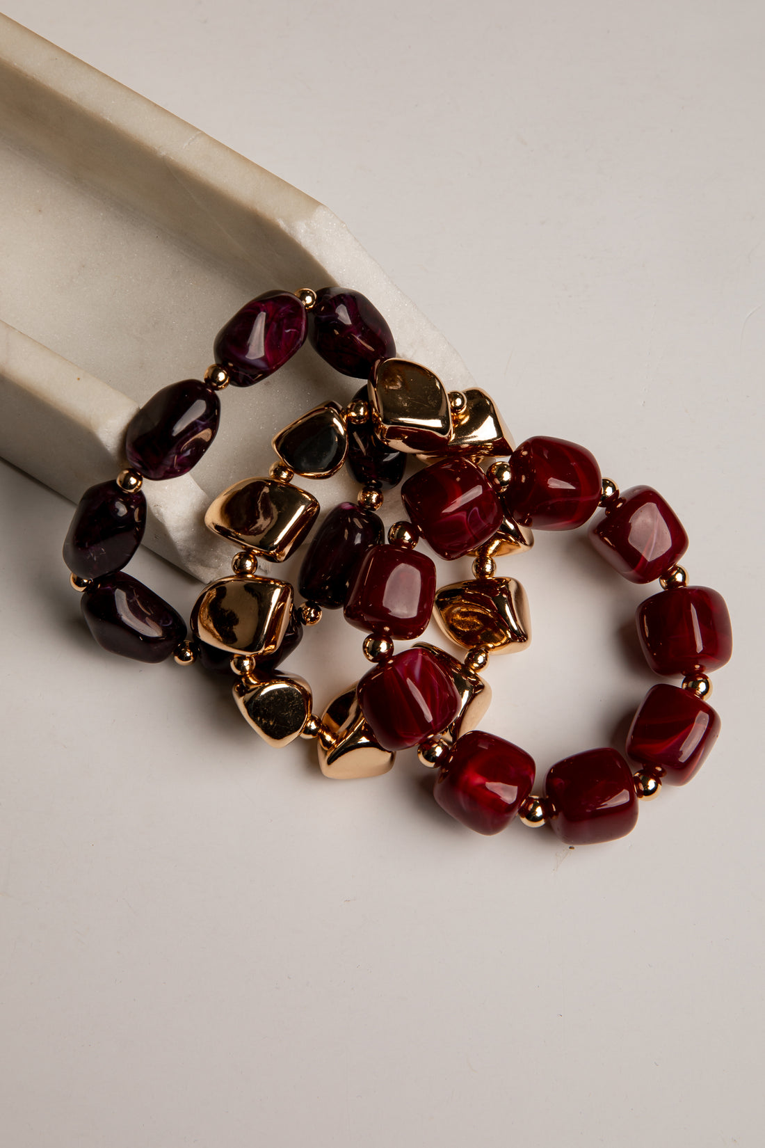 Three beaded stretch bracelets with dark red, burgundy, and black faceted beads separated by gold-toned spacers, displayed on a light beige surface.