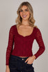 A smiling blonde woman wearing a long-sleeve burgundy lace crop top and dark blue jeans, photographed against a plain light gray studio background.