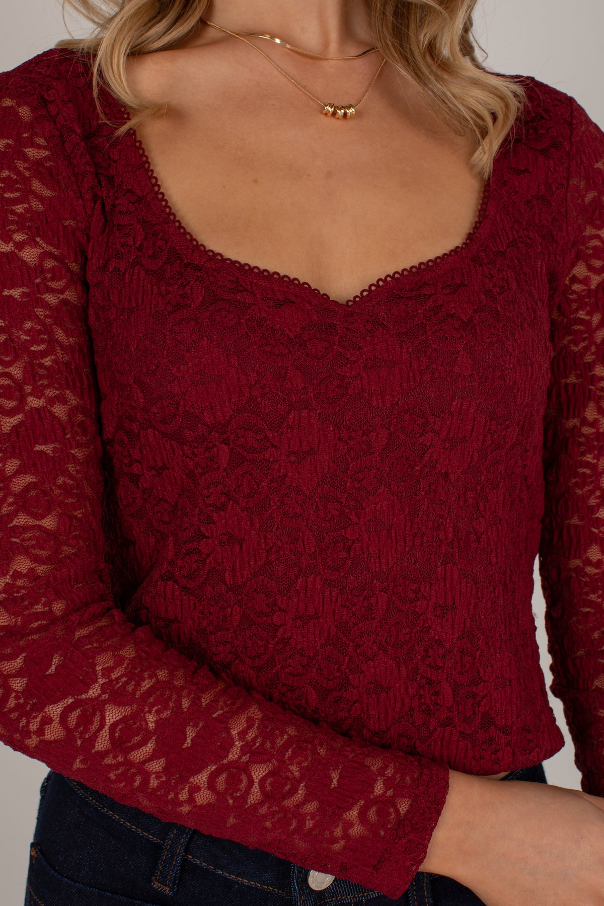 A close-up of a woman wearing a long-sleeve burgundy lace top with a scalloped scoop neckline, paired with dark blue jeans and layered gold necklaces.