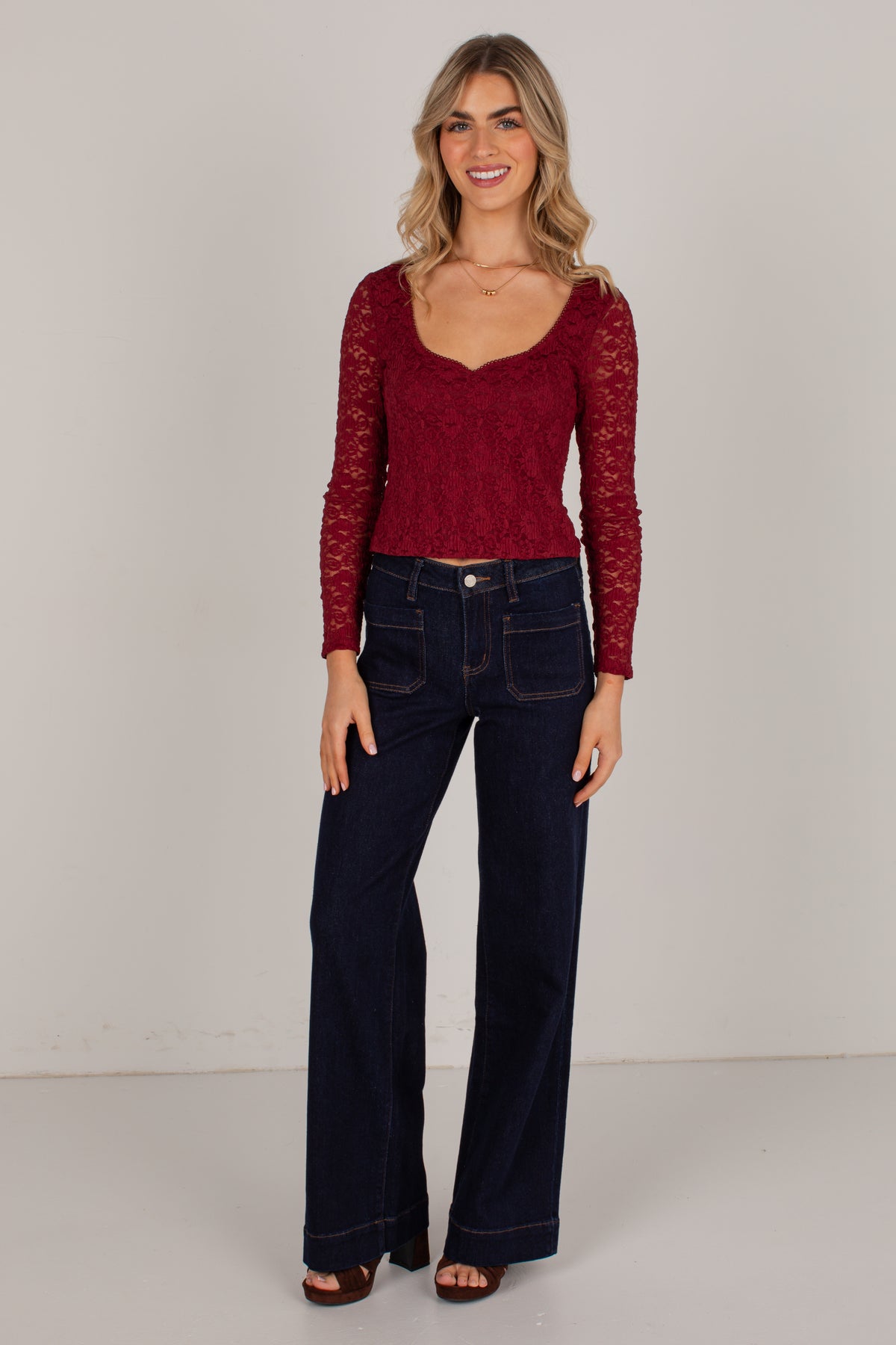 A woman modeling a burgundy lace long-sleeve scoop-neck top paired with dark blue wide-leg jeans, standing against a plain light gray studio background.