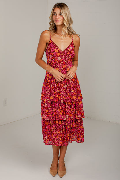 A woman modeling a tiered midi dress with a vibrant red, pink, and orange floral pattern, standing against a plain white studio background.