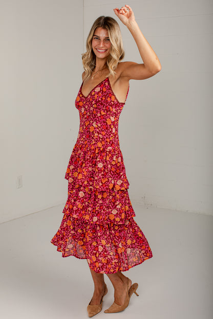 A woman modeling a sleeveless tiered midi dress with a vibrant red, orange, and pink floral print, paired with tan high heels in a bright studio setting.