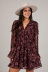 A woman wearing a dark burgundy floral mini dress with sheer long sleeves and a wide-brimmed cream hat, posing against a light gray studio background.