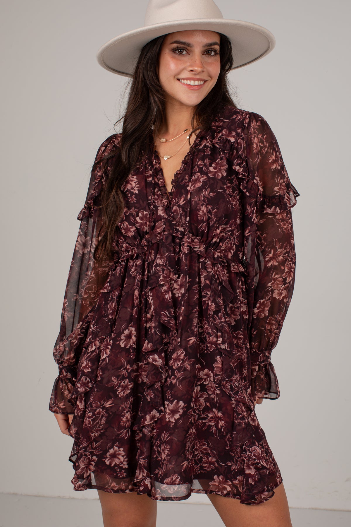 A woman wearing a dark burgundy floral long-sleeve mini dress with sheer ruffle details and a wide-brimmed cream hat, posing against a light gray studio background.