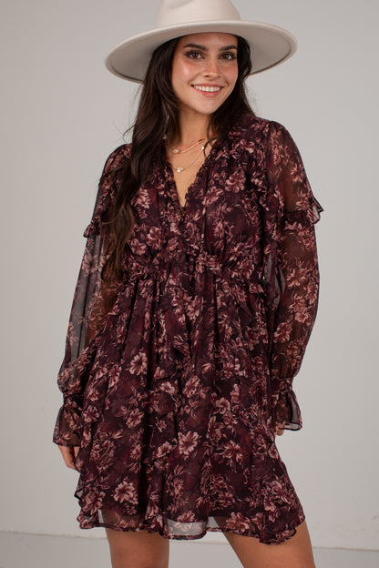 A woman wearing a dark burgundy floral long-sleeve mini dress with sheer ruffle details and a wide-brimmed cream hat, posing against a light gray studio background.