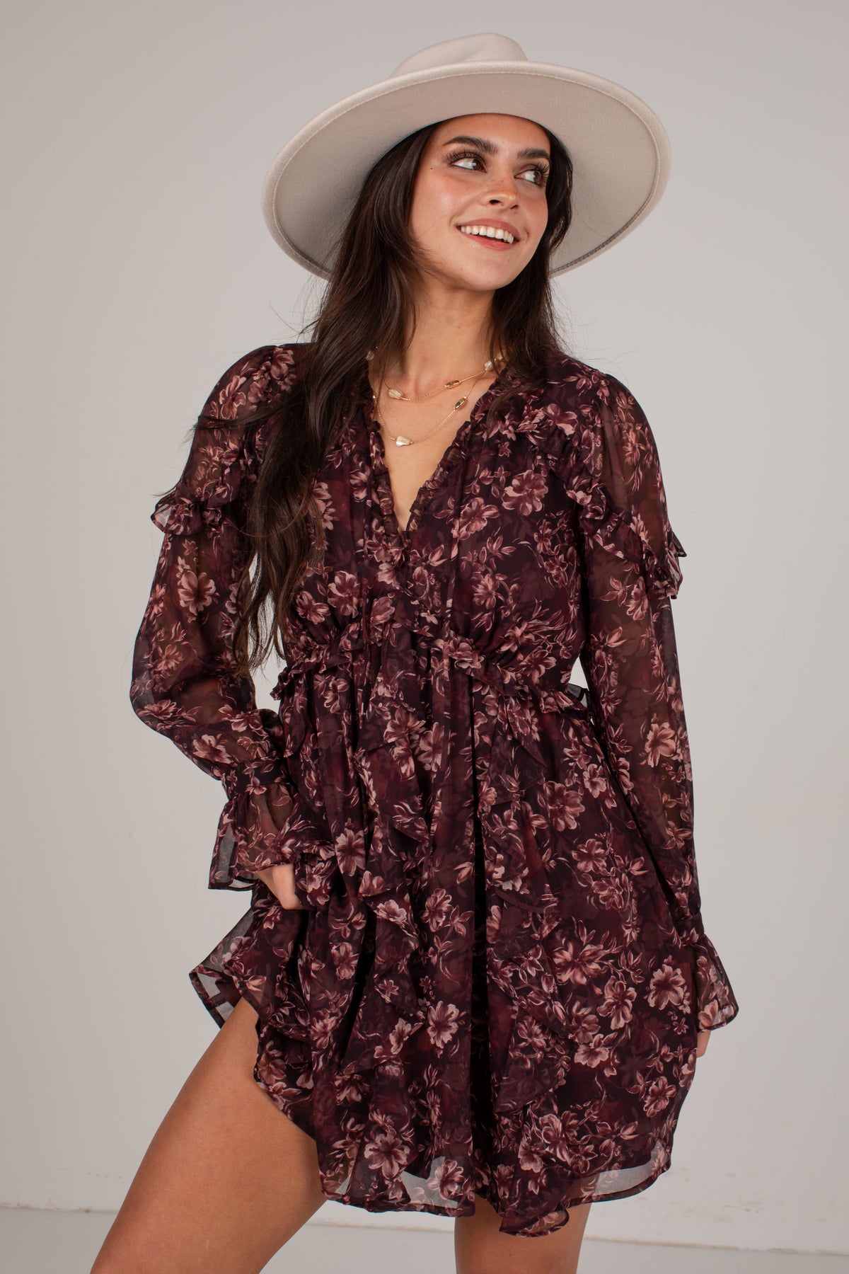 A woman wearing a dark burgundy floral long-sleeve mini dress with a ruffled V-neck and waist, paired with a wide-brimmed cream hat, posing against a light gray studio background.
