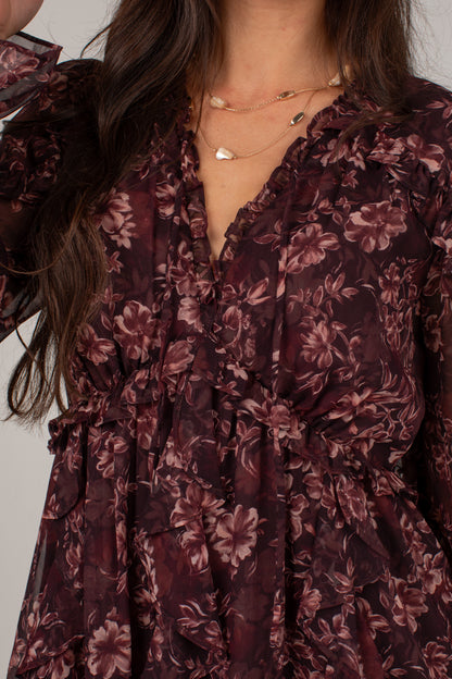 Close-up of a woman wearing a dark floral dress with a ruffled V-neckline and long sleeves, paired with layered gold necklaces.