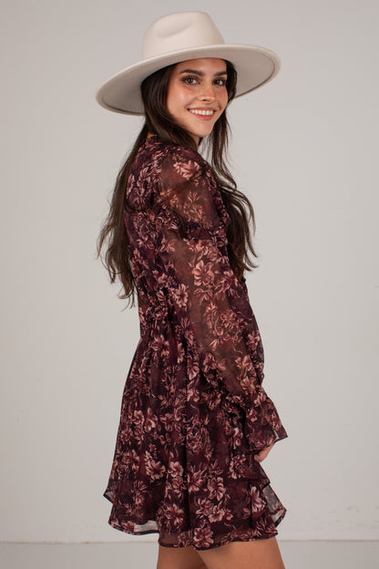 A woman wearing a sheer burgundy floral long-sleeve mini dress and a wide-brimmed cream hat, photographed against a plain light gray studio background.