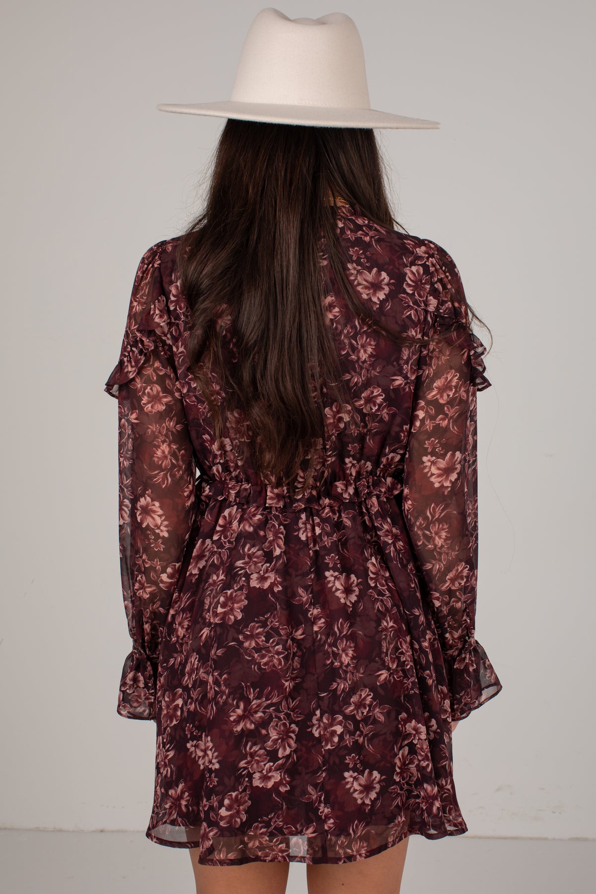 A woman viewed from behind wearing a sheer floral long-sleeve dress in dark burgundy and pink tones, paired with a wide-brimmed white hat, against a plain light gray background.