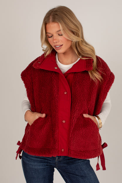 A woman wearing a red sherpa fleece button-front vest with side ties, layered over a white long-sleeve shirt and blue jeans, against a plain light gray studio background.
