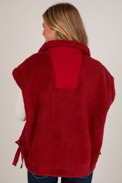A woman shown from the back wearing a red sherpa fleece vest over a white long-sleeve top and blue jeans, against a plain light gray studio background.