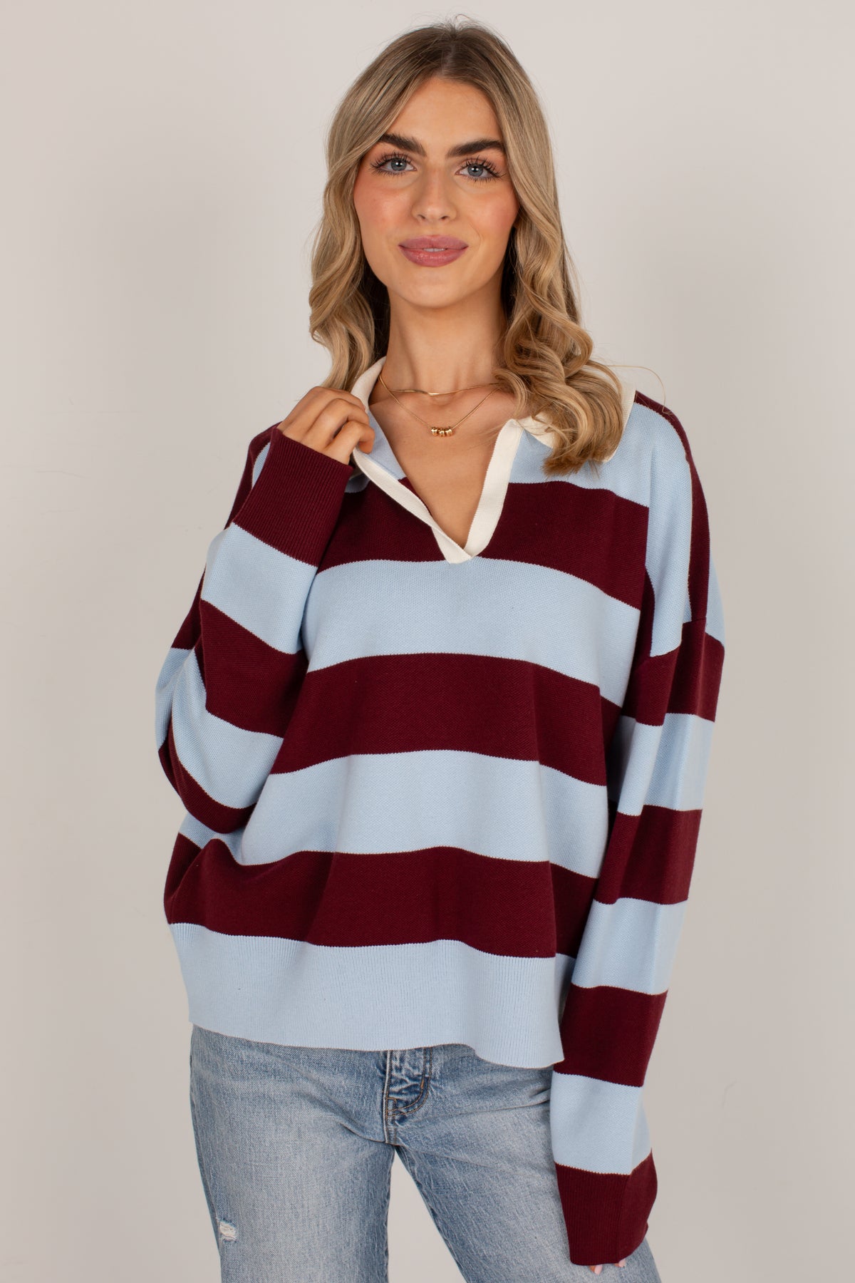 A woman wearing a light blue and burgundy striped knit sweater with a white collar detail, paired with light-wash blue jeans, against a plain light gray studio background.