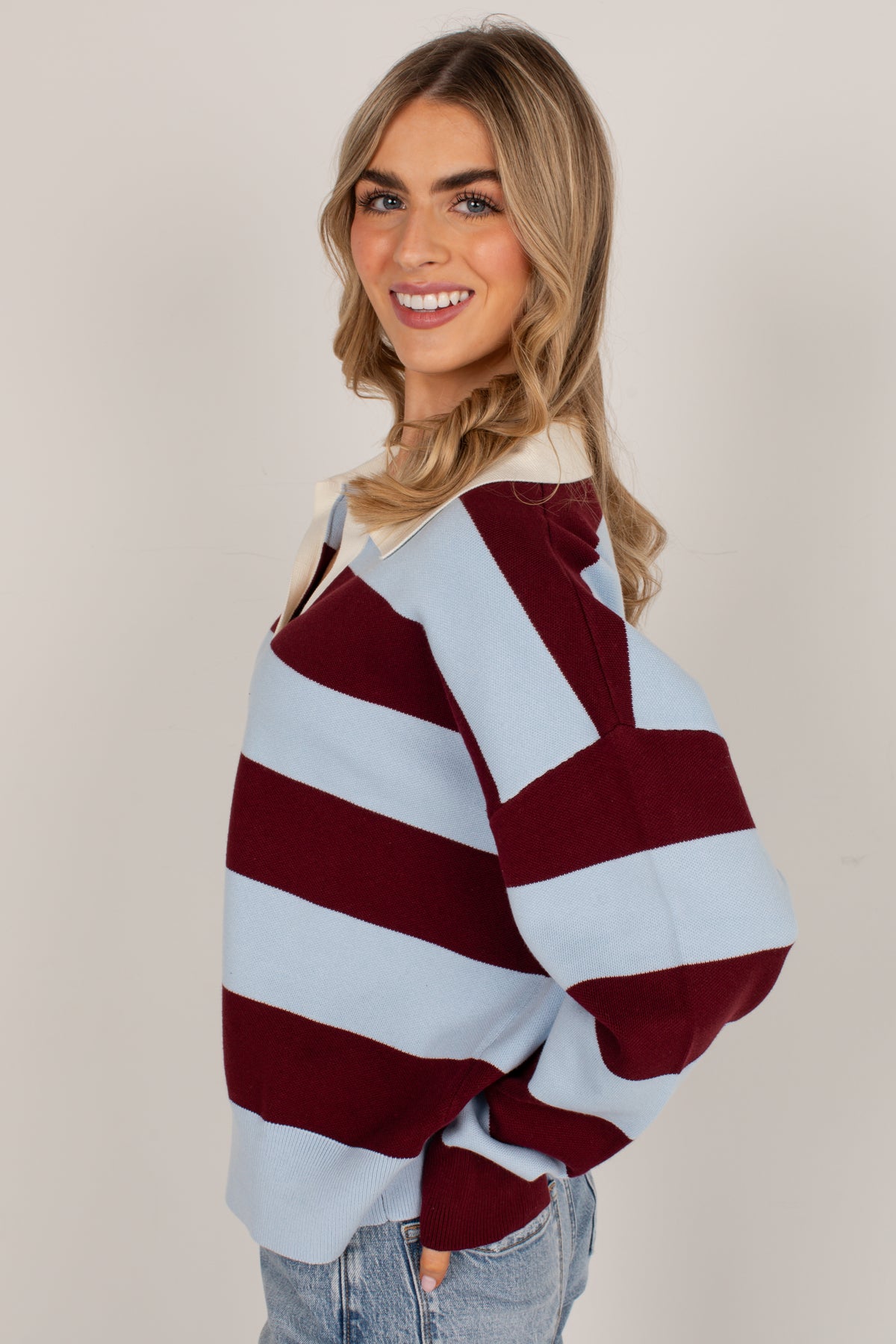 A woman modeling a loose, oversized knit sweater with wide horizontal stripes in burgundy and light blue, paired with light-wash jeans against a plain beige studio background.