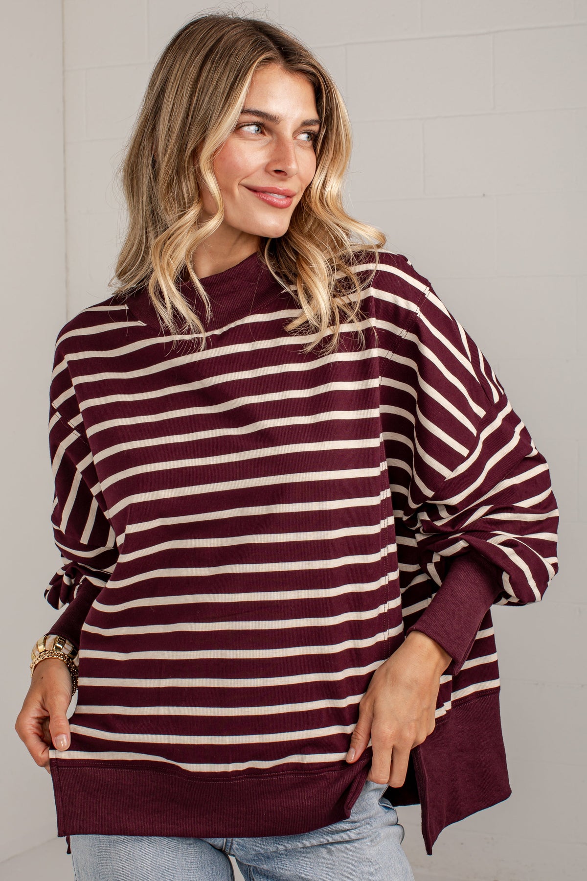 A woman wearing a loose-fitting, long-sleeve striped sweater in burgundy and cream, paired with light-wash jeans, standing against a white brick wall.
