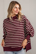 A woman wearing a loose-fitting, long-sleeve striped sweater in burgundy and cream, paired with light-wash jeans, standing against a white brick wall.