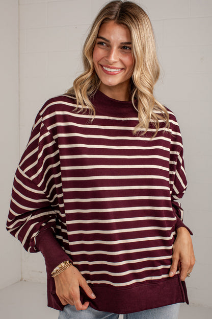 A woman wearing a burgundy and cream striped long-sleeve sweatshirt with a crew neck, paired with light blue jeans, standing against a white brick wall.
