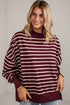 A woman wearing a burgundy and cream striped long-sleeve sweatshirt with a crew neck, paired with light blue jeans, standing against a white brick wall.