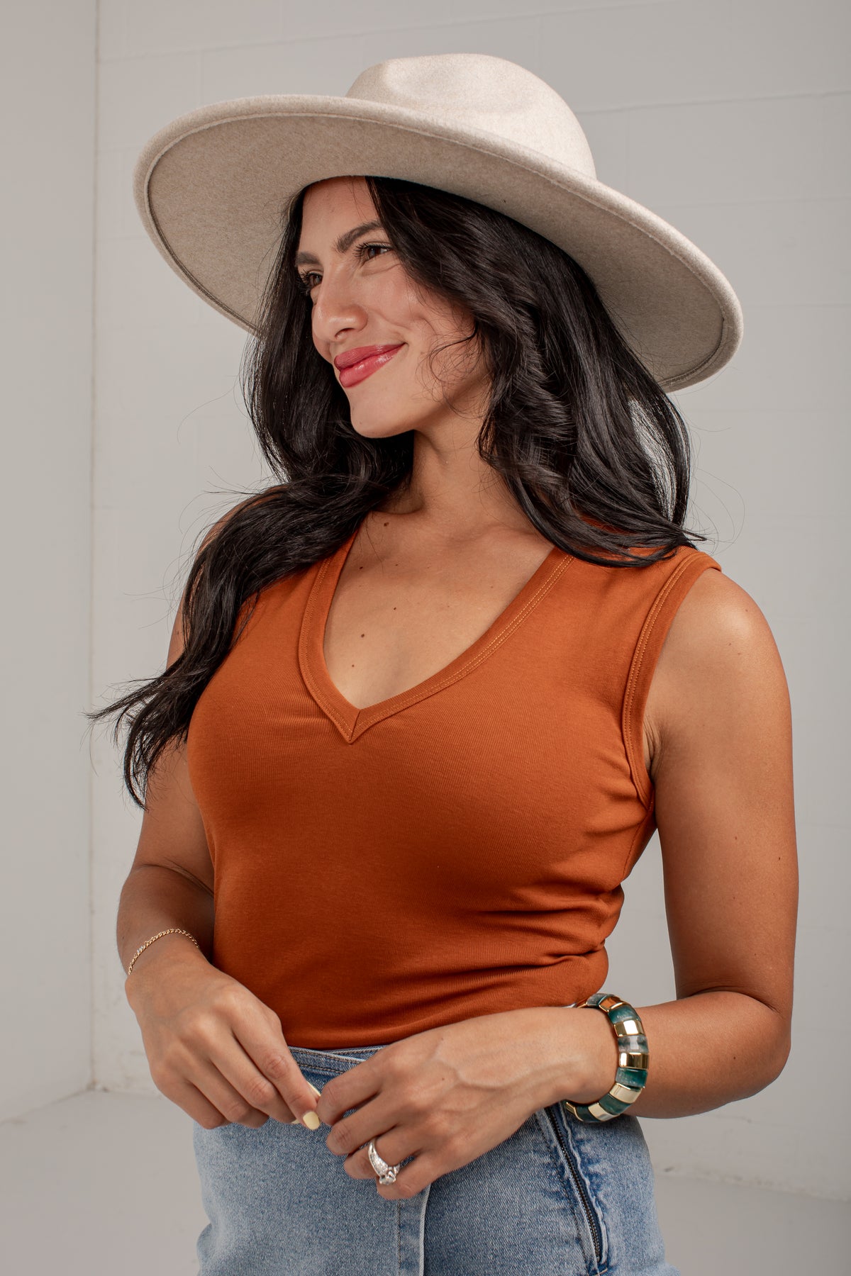 A woman wearing a rust-orange sleeveless V-neck top, a wide-brimmed cream hat, and light-wash blue jeans, standing against a light gray wall.