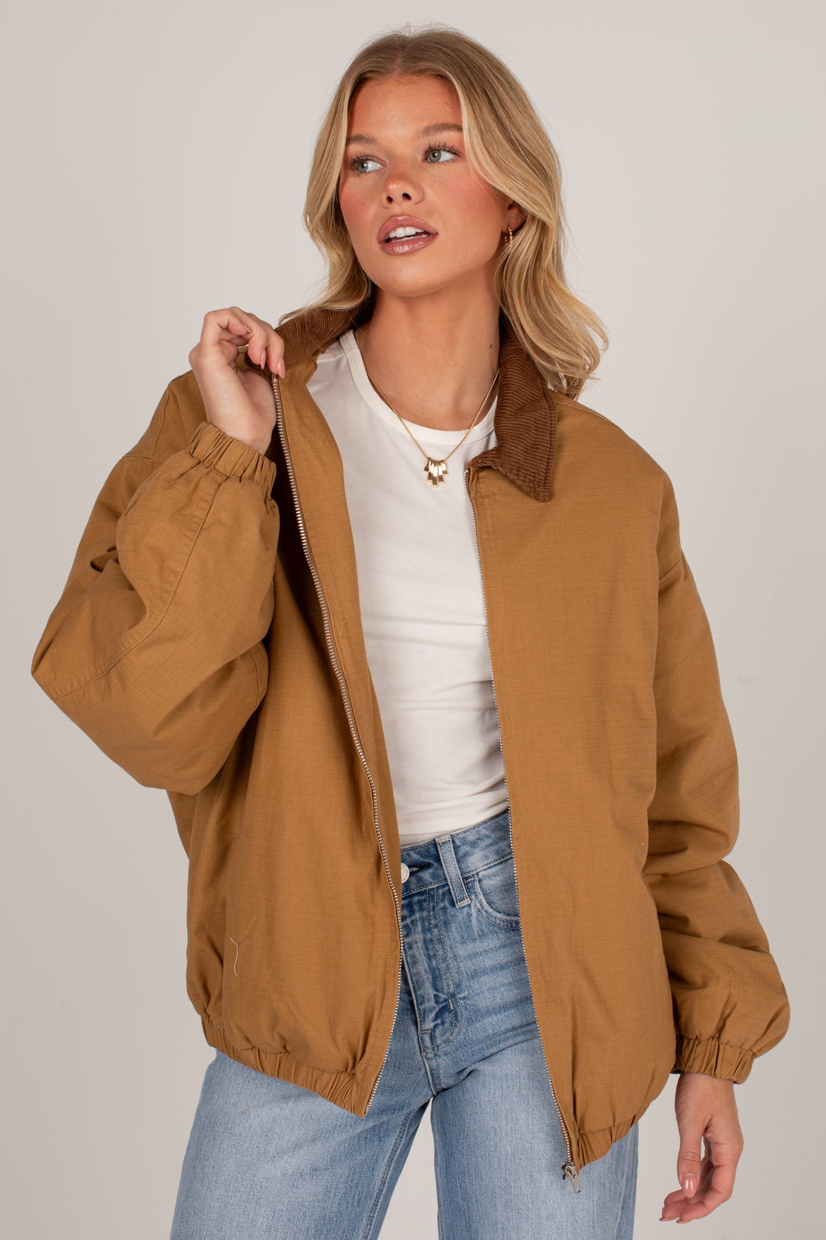 A woman wearing a tan utility jacket with a brown collar, styled with a white top and blue jeans, posing against a light gray studio background.