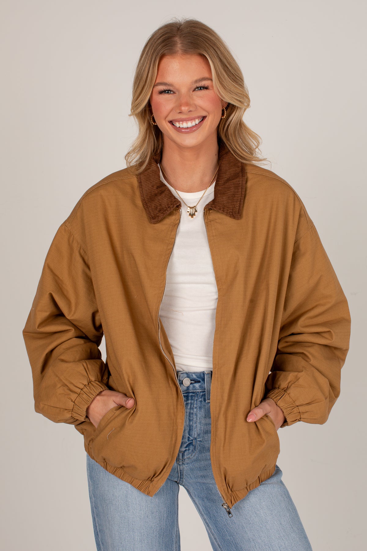 A smiling blonde woman wearing a tan oversized zip-front jacket with a brown collar, layered over a white top and blue jeans, photographed against a plain light gray studio background.