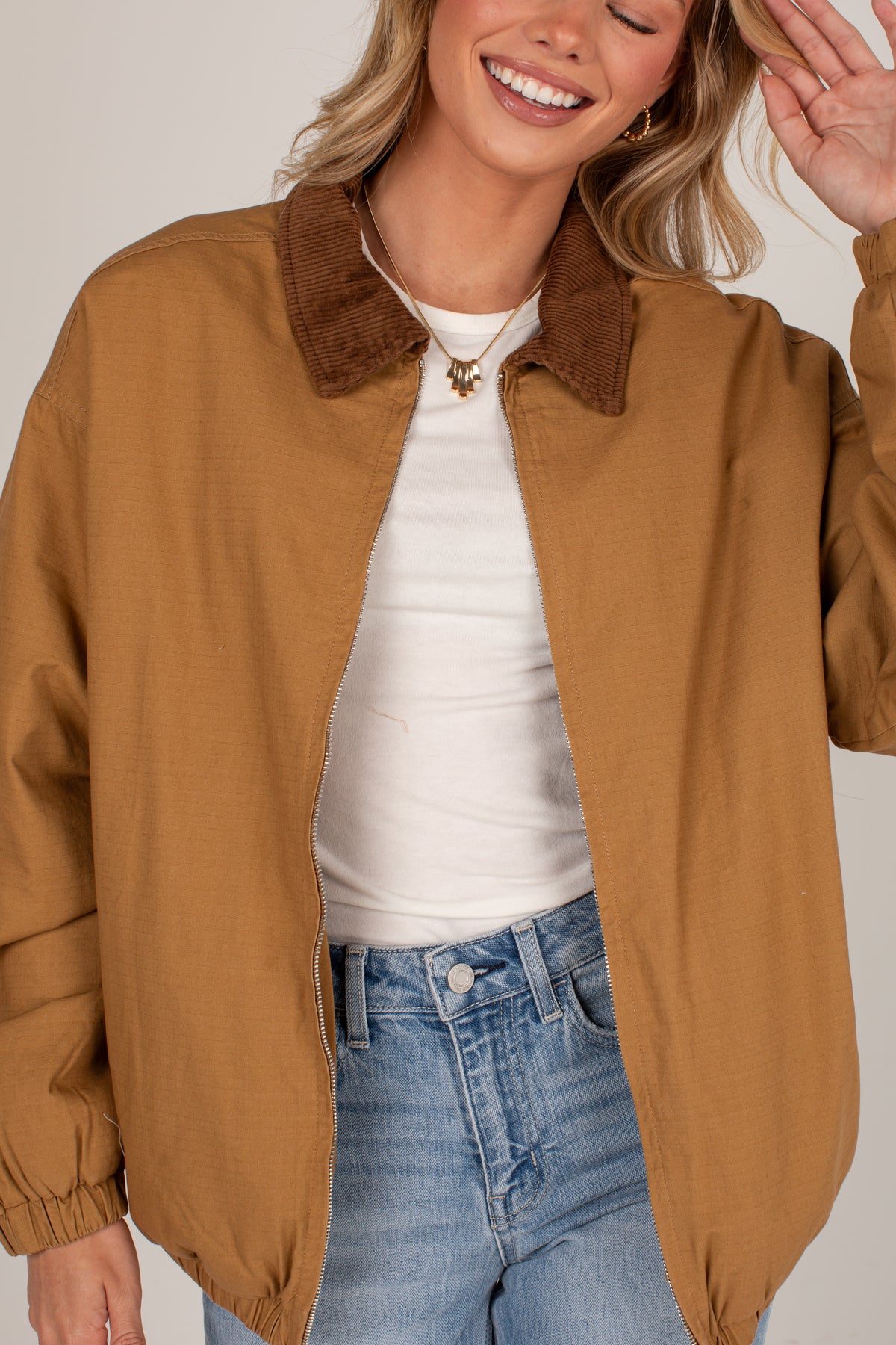A woman wearing a tan-brown jacket with a brown corduroy collar, layered over a white t-shirt and blue jeans, against a light beige background.