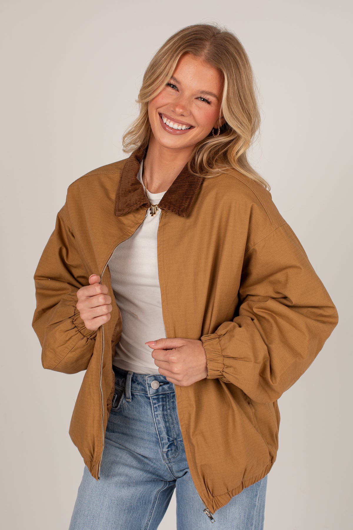 A smiling blonde woman models a tan utility-style jacket with a brown corduroy collar, worn over a white top and blue jeans, against a plain light gray studio background.