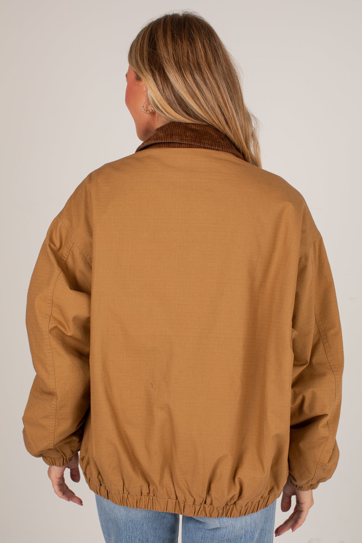 A woman viewed from behind wearing a tan utility jacket with a brown corduroy collar and elastic cuffs, paired with light blue jeans against a plain light gray background.