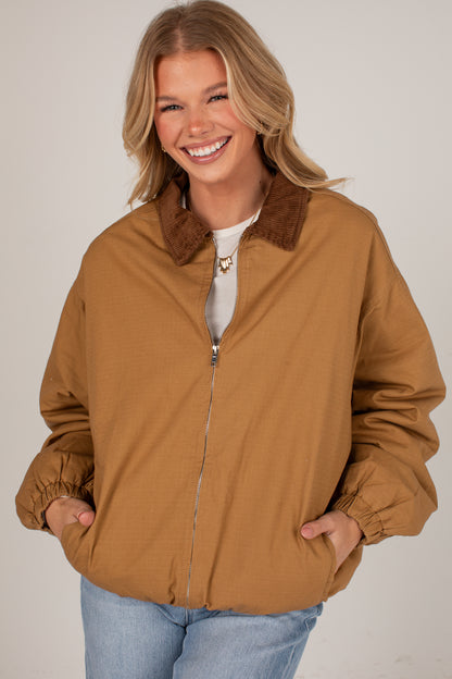 A smiling blonde woman wearing a tan zip-up jacket with a brown faux-fur collar, layered over a white top and blue jeans, against a plain light gray studio background.