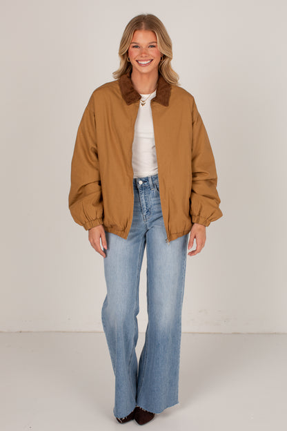 A woman modeling a tan utility jacket with a brown corduroy collar, worn over a white top and light blue wide-leg jeans, against a plain off-white studio background.