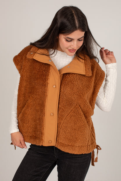 A woman wearing a brown sherpa fleece vest with tan leather trim over a white long-sleeve top and black pants, photographed against a light gray studio background.