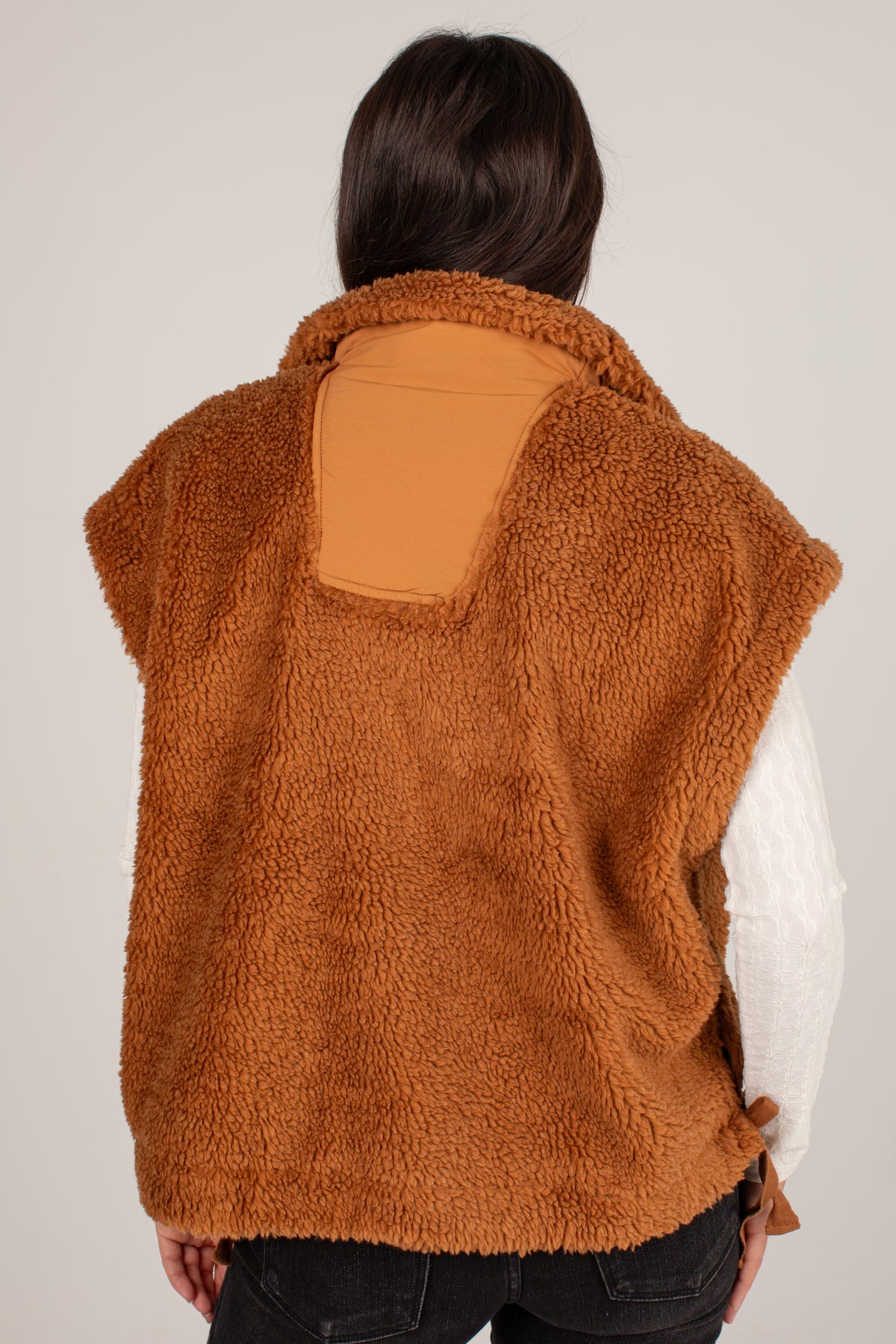 A person shown from the back wearing a brown sherpa fleece vest over a white long-sleeve shirt and dark jeans, against a plain light gray studio background.