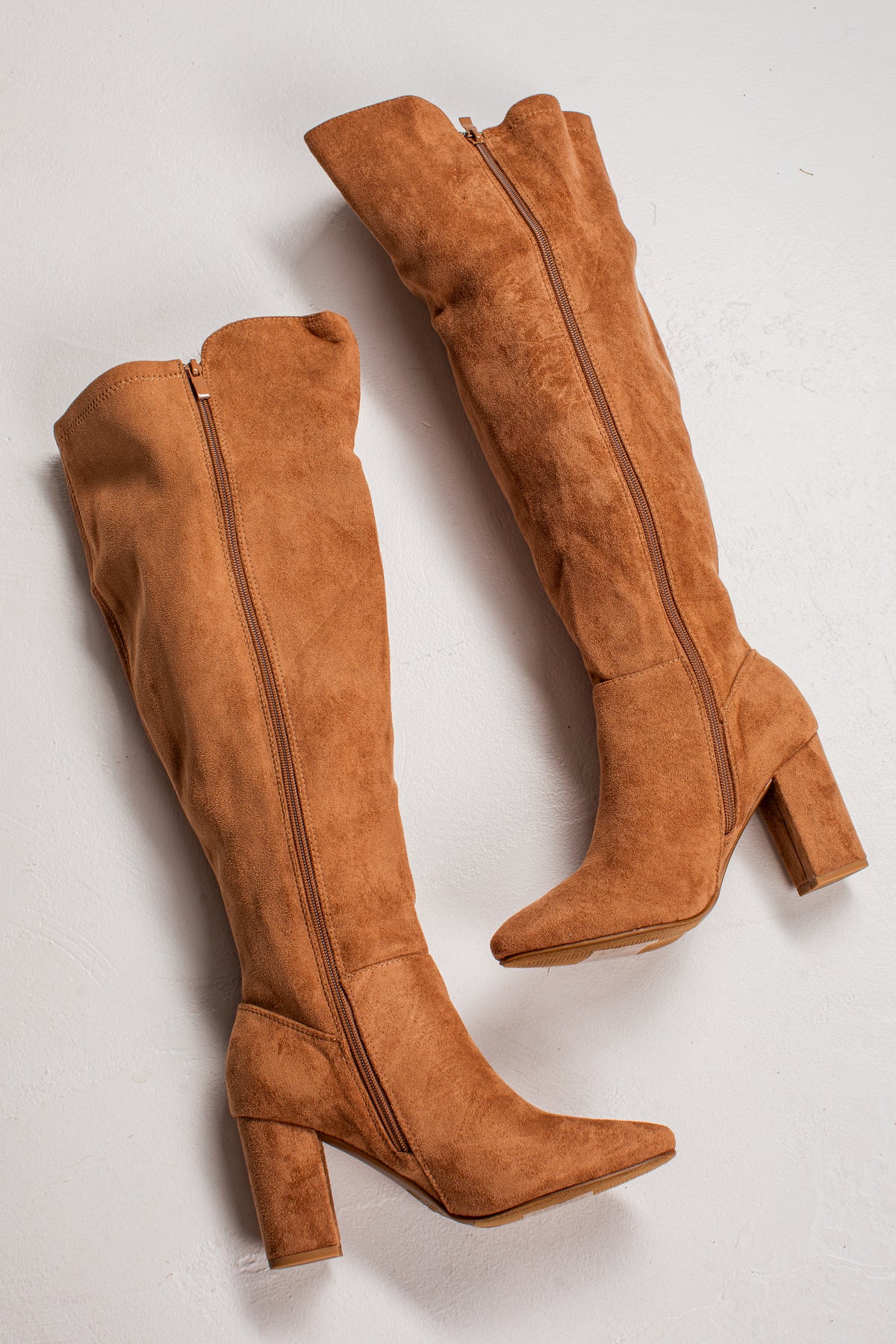 A pair of knee-high suede boots in a warm camel brown color, featuring a side zipper, pointed toes, and block heels, displayed on a light gray background.