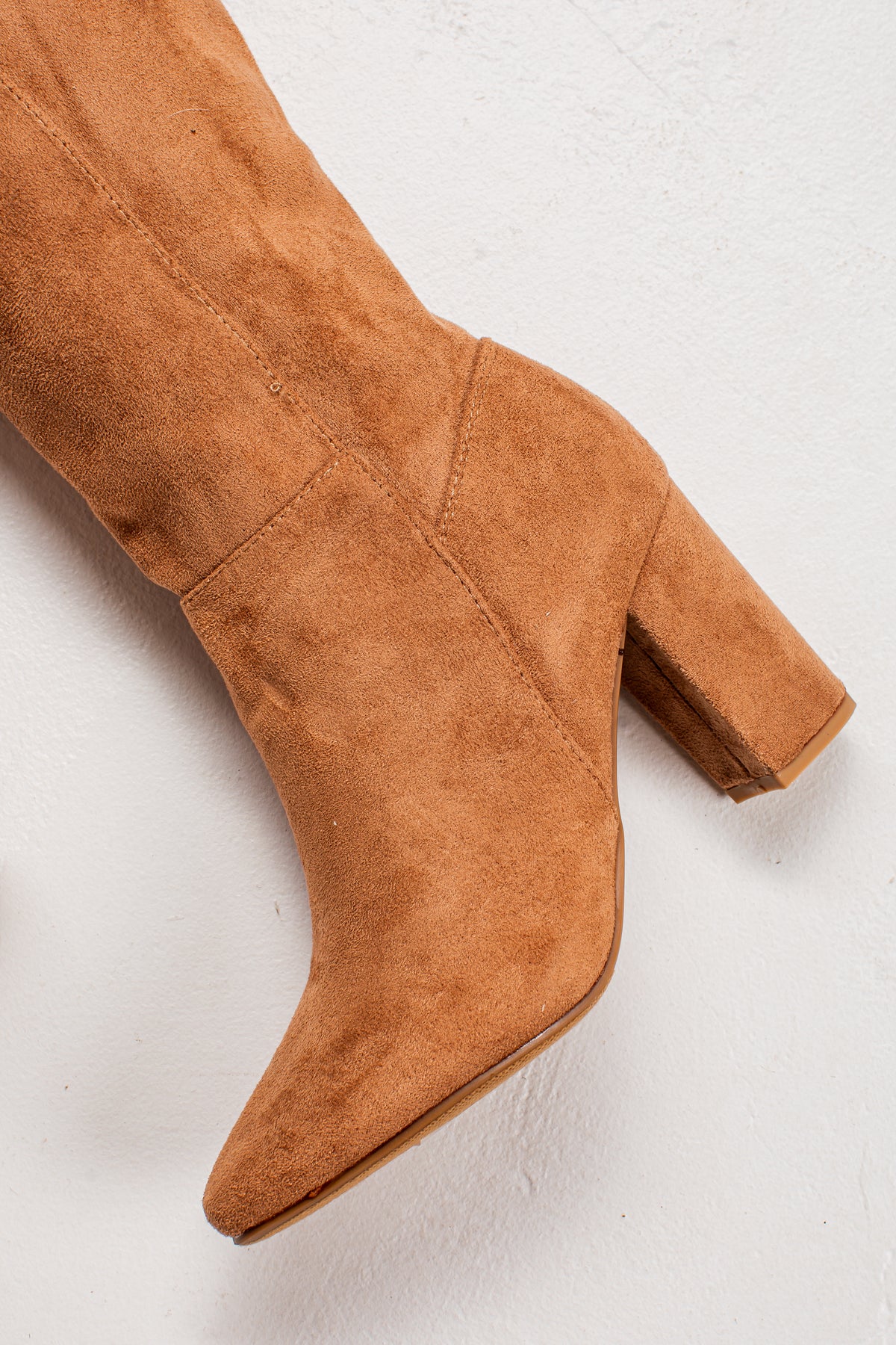 A close-up of a tan suede knee-high boot with a block heel and pointed toe, shown against a light textured background.